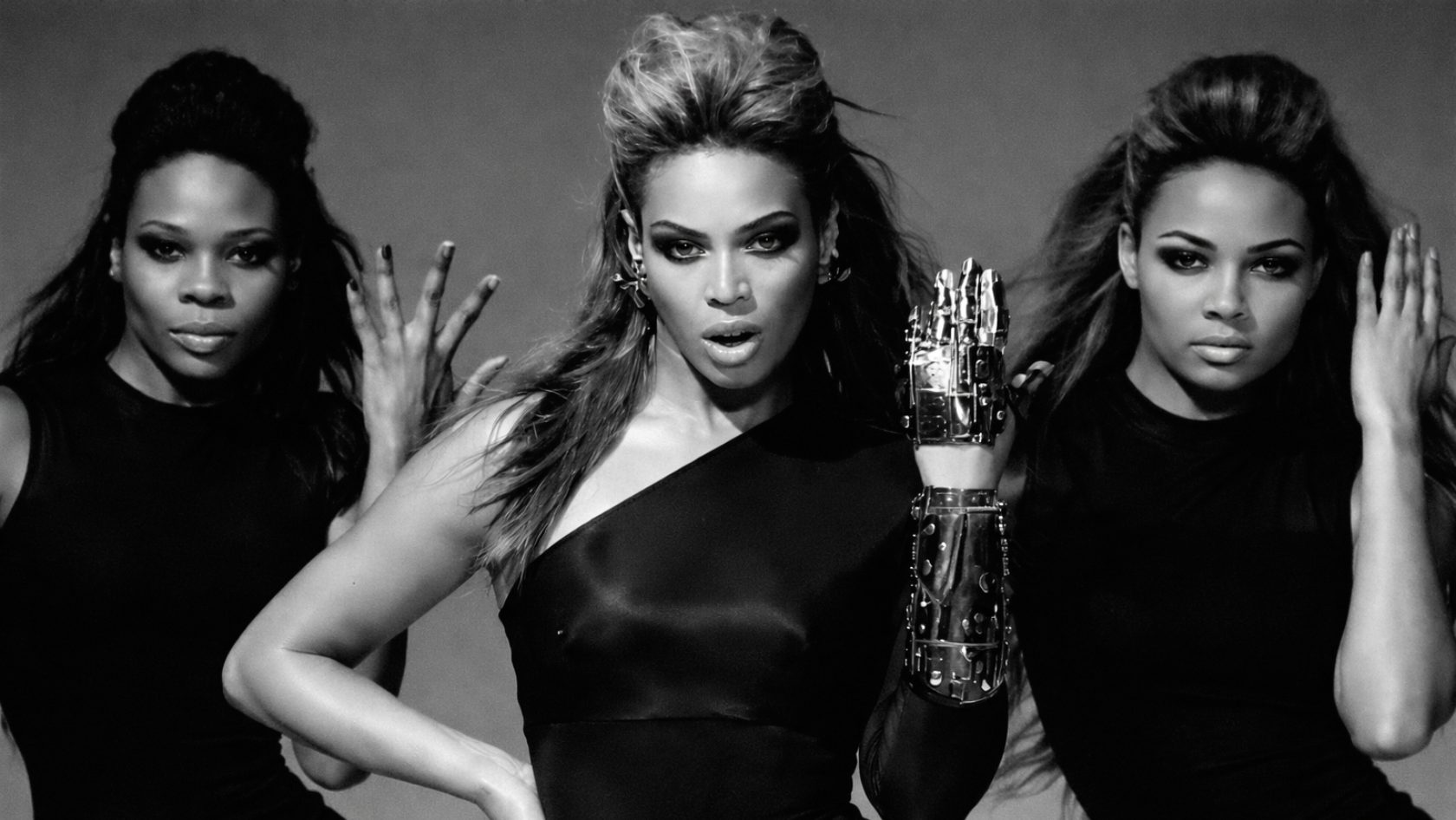 Black and white editorial photograph of Beyoncé in the center, flanked by two dancers. Beyoncé, with a high coif hairstyle, wears a sleek black one-shoulder dress and displays an intricate, chrome robotic gauntlet and sleeve on her left arm. The dancers are in matching black outfits with powerful hand poses. This image is an iconic representation of her "Sasha Fierce" era identity.